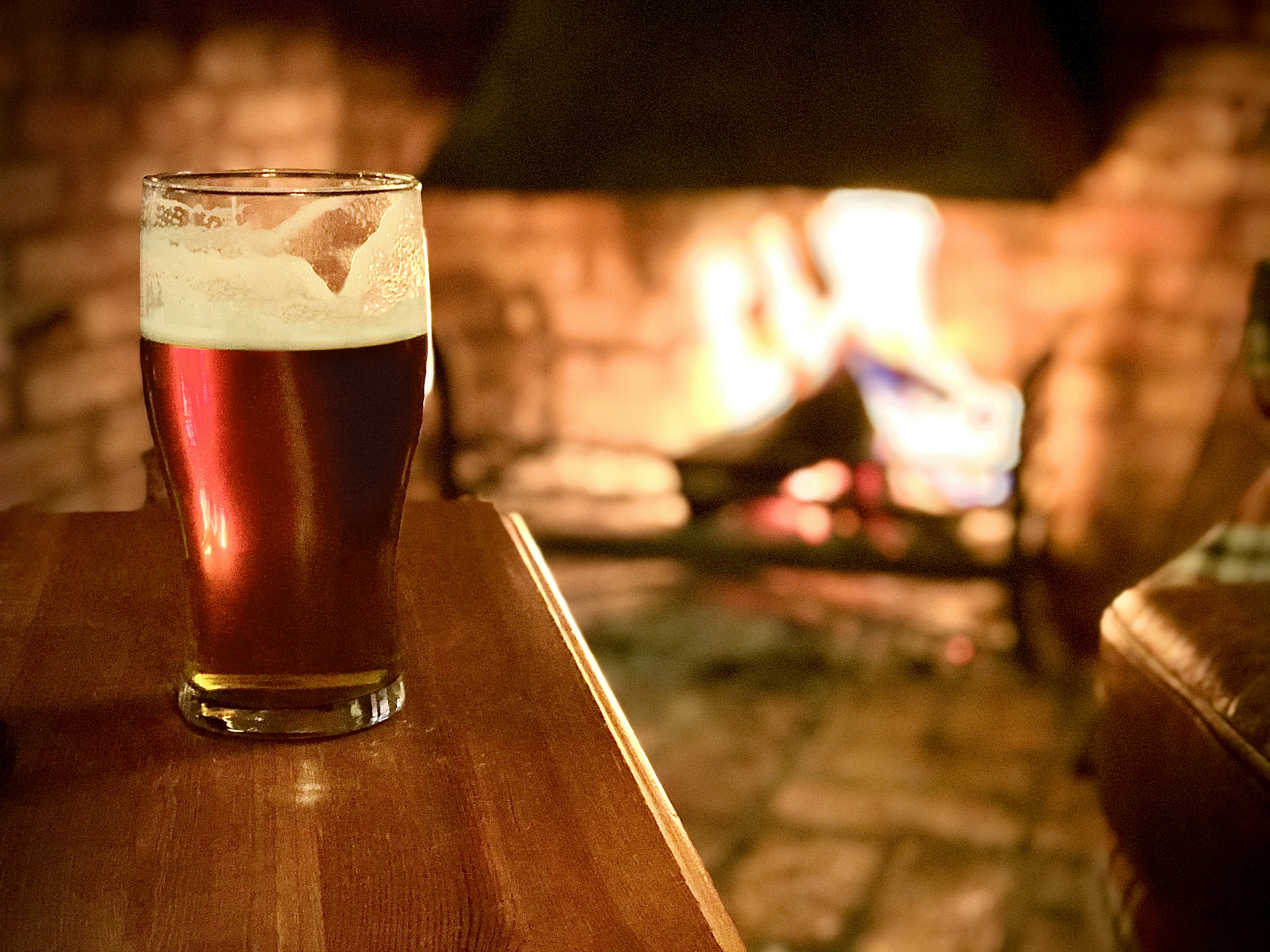 A pint of beer on a wooden table beside a glowing fireplace in a cosy traditional pub